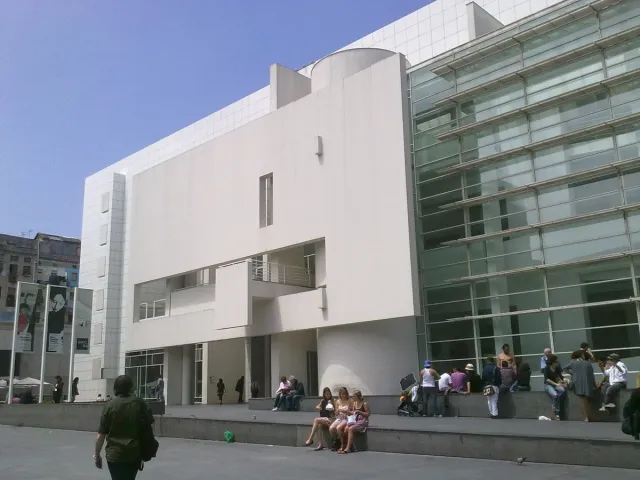 מבנה לבן גדול עם חזית זכוכית ואנשים יושבים על הכיכר שלפניו