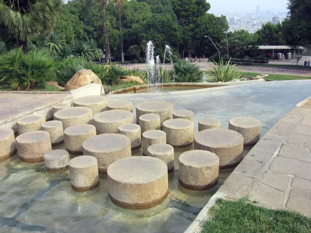 גלילי אבן בתוך בריכה רדודה עם סילוני מים וגן דקלים מעליה