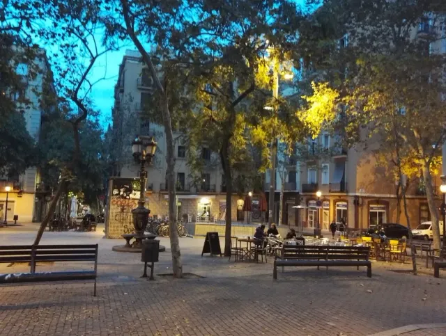 כיכר מרוצפת עם עצים, פנס ברזל, ספסלים ושולחנות מרפסת בשעת ערב