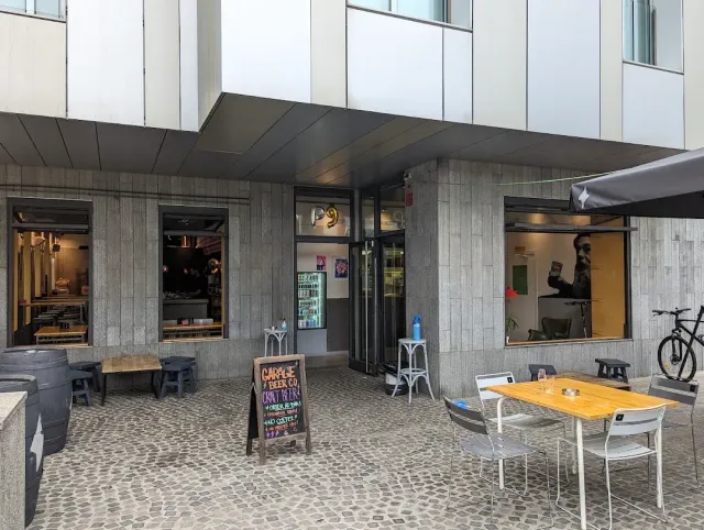 כניסה בבניין בטון עם שלט גיר, שולחנות חוץ ואופניים על רחבה מרוצפת