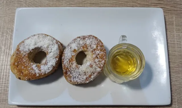 שני מאפים טבעתיים מפוזרים באבקת סוכר וכוסית נוזל זהוב, בצלחת לבנה