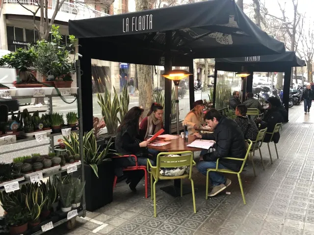 מרפסת רחוב בסוכך שחור עם כיסאות ירוקים, סועדים וחנות צמחים לצד