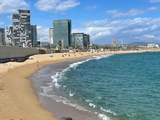 גלים קטנים נשברים על חול רטוב, ומגדלי זכוכית בקצה הטיילת