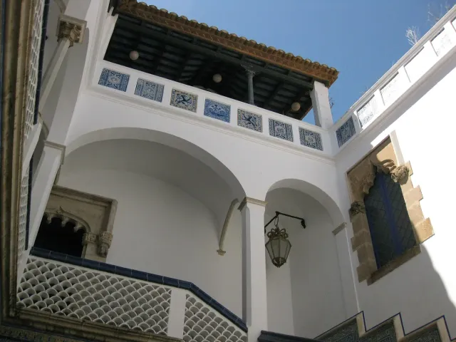 חצר לבנה עם קשתות, אריחי קרמיקה בכחול לבן ופנס תלוי מול שמים כחולים